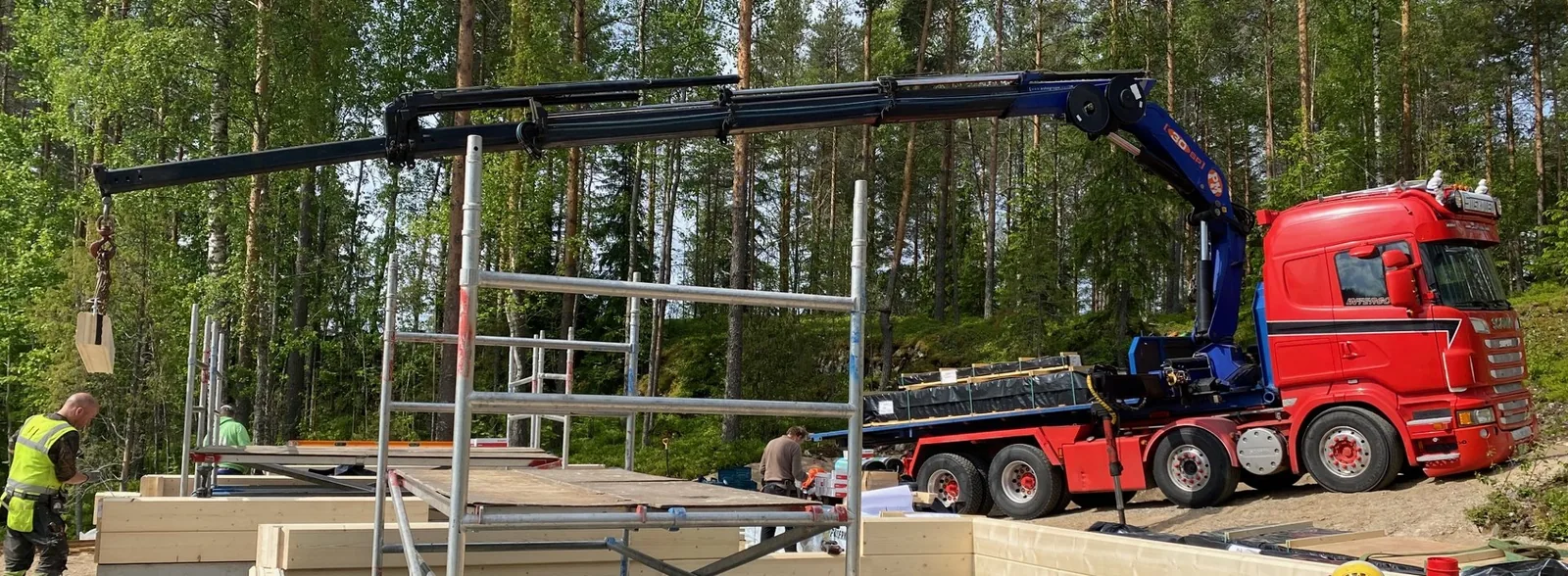 Nosturin puomi hirsikehikon ja telineiden yllä, koukussa hirsi.